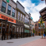A street view of a restaurant named "Mexican Kitchen" housed in a charming brick building. The area features similar style buildings, trees, and a clear sky, enhanced by premium electric streetlights that illuminate the scene beautifully.