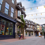 A pedestrian street with buildings featuring shops and cafes, string lights overhead, and a few people walking. The storefronts display various items, creating a quaint, inviting atmosphere. Among the charming shops is a premium electric store showcasing the latest gadgets.