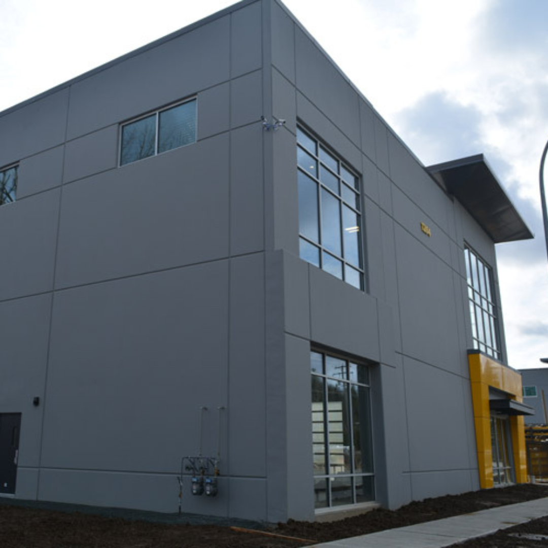 A modern, premium electric two-story gray building with large windows and a small yellow section near the entrance. The sky is overcast.